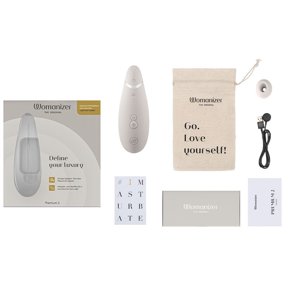 Tillbehör till Womanizer Premium 2 Warm Gray. Laddare till lufttrycksvibrator och påse med text &quot;Go. Love Yourself!&quot; och vit låda.