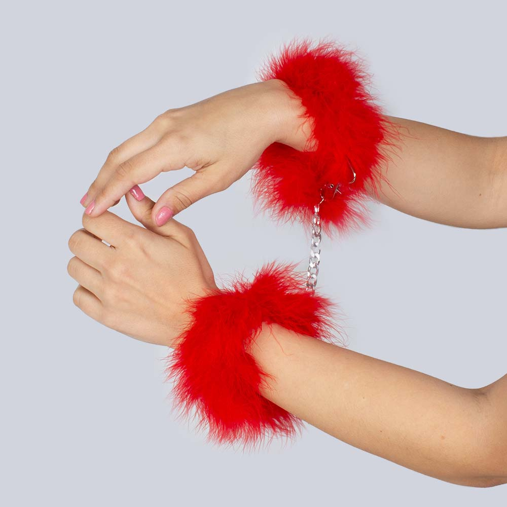 Två händer iförda Secret Play Fuzzy Handcuffs Red. Nagellack på naglarna. 