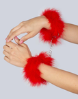 Två händer iförda Secret Play Fuzzy Handcuffs Red. Nagellack på naglarna. 